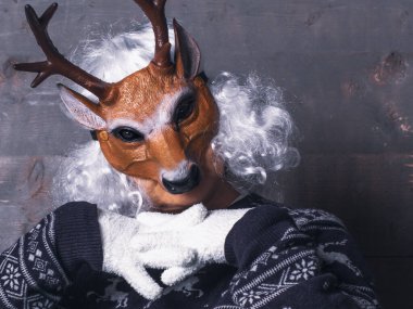 Geyik maskeli ve kışlık elbiseli bir kadın rahat bir pozisyonda oturur. Kırsal stil, kaba ahşap zemin, havalı ton.