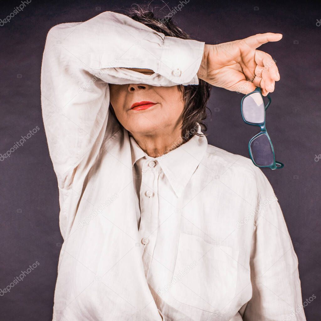 Mature woman in a white linen shirt, covers her face with her arm and holding glasses in her hand.