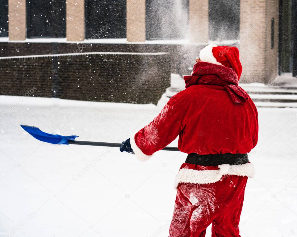 Santa Claus in a traditional costume with a shovel removes snow on a snowy street.