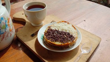 A mini photo of a martabak with a chocolate topping and grated cheese found a cup is hot, a knife, a fork and cutlery