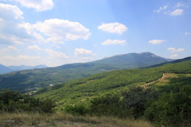Yaz dağı manzarası. Dağ yamacındaki yeşil orman güneş tarafından aydınlatıldı. Mavi gökyüzü ve beyaz bulutlarla aydınlık bir gün..