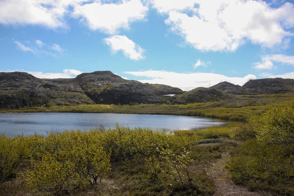 Summer landscape with the lake on a sunny day. The hills with snow are illuminated by the sun. A walking path runs between green bushes. Bright green grass surrounds the lake.