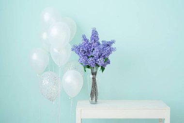 Masadaki yüksek vazoda mavi bir duvarın arkasında duran leylak demeti. Tatil için beyaz balonlar.
