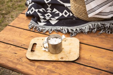 Parktaki ahşap bir masada marşmelovlu bir kupa kakao. Ekose, sepet ve piknik kupası..
