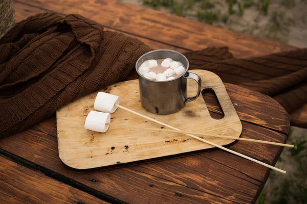 Travel tin mug with cocoa and marshmallows and marshmallows on sticks for making smore. Wooden table in a camp or park. Dessert on a hike.