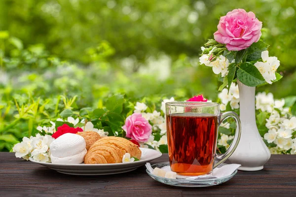 杯子里有茶和糖 羊角面包 棉花糖 白花瓶和一束茉莉花枝条 还有一朵玫瑰花在开着花的灌木丛中的木桌上 户外野餐早午餐选定重点 — 图库照片
