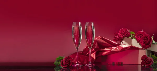 Luxurious glasses with sparkling wine open box with glitter brocade surrounded by selected roses on a mirror surface on a red background. Valentine\'s day or romantic evening invitation