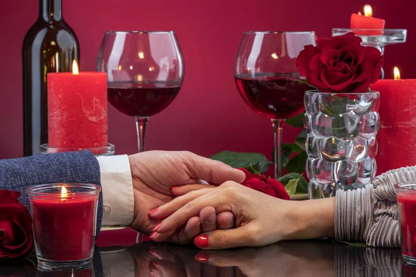 Male and female hands are displayed in a mirror surface among melting candles, red roses, glasses of wine.