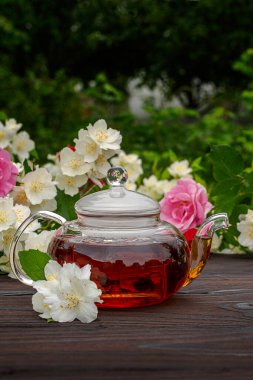 Cam buğulanmış çaydanlık, çiçek açan gül ve yasemin çalılarının arasında, ahşap bir masada. Açık hava, piknik, kahvaltı. Yeşil yaprakların arkaplanı bulanık.