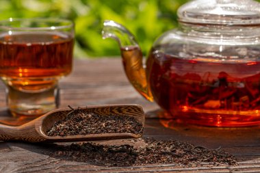 Kırpılmış cam, buğulanmış çaydanlık manzarası, zarif bir fincan. Çaylı bambu kaşığı, ahşap masa. Açık hava, piknik, kahvaltı. Bulanık çiçekli arkaplan.