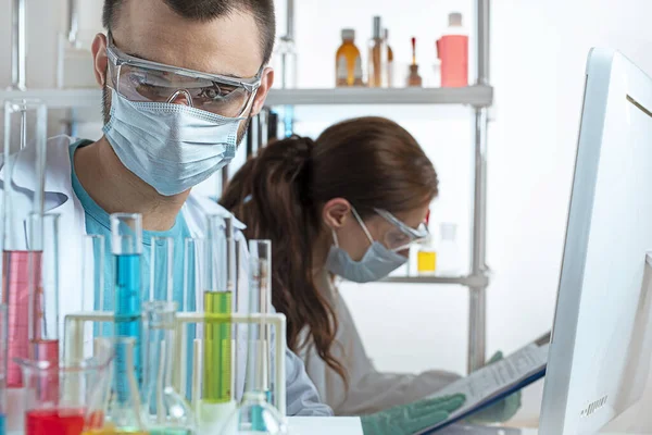 Cropped view of two young scientists in protective equipment in a laboratory. Selected focus on man looking at beakers. Woman working with data in computer