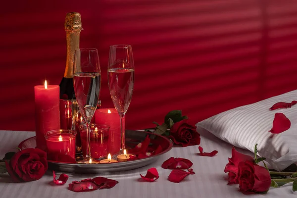 A fragment of a bed in a luxurious hotel or at home with a tray with candles, a bottle in an ice bucket, glasses of wine, a decoration of red roses and petals. White linens, red wall with silhouette or shadow. Honeymoon, or Valentine's Day