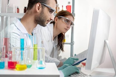 İki genç bilimadamı, laboratuvar ekipmanları ve tıbbi camlar arasında bir bilgisayardaki veriye bakıp çalışırlar.