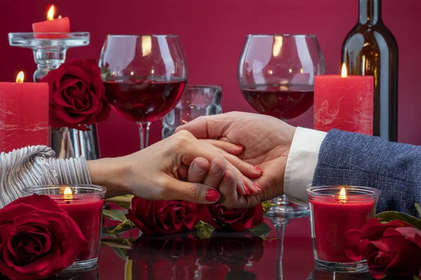 Elegant male and female hands are displayed in a mirror surface among melting candles, red roses, glasses of wine. Gala evening.