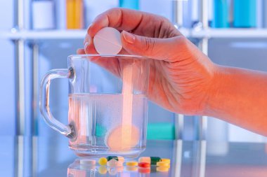 Close-up view of hand throwing dissolving effervescent tablet in glass cup and scattered tablets, pills and drugs in the hospital. Glass tabletop with reflections in sterile room. Close up view clipart