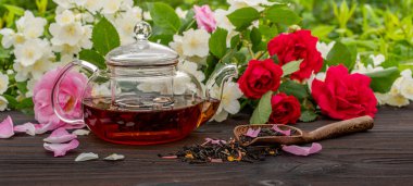 Buharda pişirilmiş cam çaydanlık ve bambu kaşıkla çiçek açan gül ve yasemin çalılarının arasında çay. Açık hava, piknik, kahvaltı. Bulanık çiçekli arkaplan.