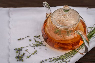 Ahşap arka planda beyaz bir masa örtüsü üzerinde lavanta çayı ile buharlı cam çaydanlık..