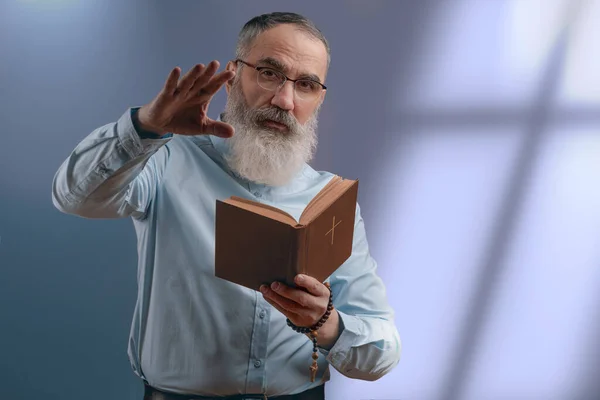Senior charismatic man with a gray beard in a light shirt holding a book Holi Bible and gestures with his hand. Light from window on gray wall on background