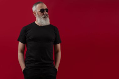 Siyah tişört giyen, gri sakallı, yetişkin ve yakışıklı bir erkek model. Ön manzara. Logonuz ya da tasarımınız için koyu kırmızı arkaplanda boşluk oluşturun