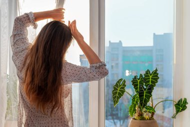 Uzun kahverengi saçlı, günlük gömlekli bir kadın perdeyi çekiyor, yüzünü güneş ışınlarından gelen eliyle kapatıyor, yaşam tarzıyla. Pencere pervazında kapalı bir bitki var. Şehrin ön cephesi silueti