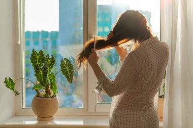 Şeffaf gömlekli bir kadın, uzun kahverengi saçlarını bir pencerenin önünde, güneş ışığıyla tarıyor. Sabah güzellik programı, yaşam tarzı. Pencere pervazında kapalı bir bitki var. Şehrin ön cephesi silueti