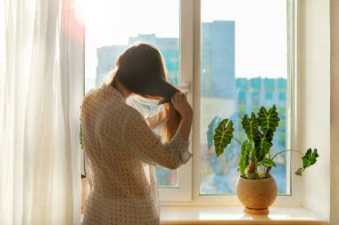 Şeffaf gömlekli bir kadın, uzun kahverengi saçlarını bir pencerenin önünde, güneş ışığıyla tarıyor. Sabah güzellik programı, yaşam tarzı. Pencere pervazında kapalı bir bitki var. Şehrin ön cephesi silueti
