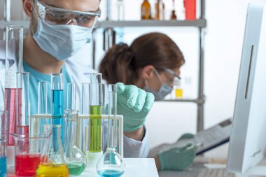 Cropped view of two young scientists in protective equipment in a laboratory. A man looking at beakers, a woman working with data in a computer. Selected focus on male hand