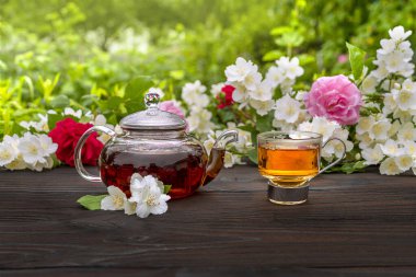 Buharda pişirilmiş bir çaydanlık ve çiçek açan gül ve yasemin çalılarının arasında zarif bir cam bardak. Açık hava, piknik, kahvaltı. Yeşil yaprakların arkaplanı bulanık.