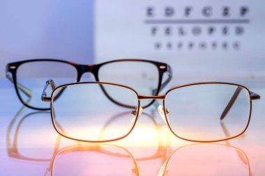 Eyeglasses with prescription lenses on a backlit glass table against a visual test with letters in blur background. Vision correction and ophthalmology concept. Selected focus clipart