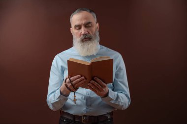 Karizmatik, gri sakallı, elinde kitap tutan bir dindar adam. Elinde haç olan, kahverengi bir arka planı olan bir haç.