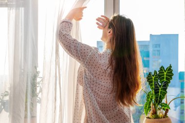 Perdeyi geri çeken şeffaf gömlekli bir kadın, elini güneş ışınlarından yüzünü kapattı, yaşam tarzı. Pencere pervazında kapalı bir bitki var. Şehrin ön cephesi silueti