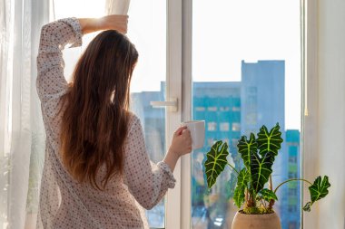 Rahat gömlekli, uzun kahverengi saçlı, bir fincan çay ya da kahve içmiş bir kadının pencereden dışarı bakıp perdeyi çekişi. Yaşam tarzı, doğal güneş ışığı..