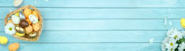Paskalya tebrik kartı, takvim, poster, afiş. Çikolata, altın, dekoratif yumurta, papatya çiçeği, açık mavi tahta üzerinde kalp şeklinde sepet.