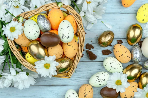 Paskalya tebrik kartı, takvim, poster, afiş. Çikolata, altın, dekoratif yumurta, papatya çiçeği, açık mavi tahta üzerinde kalp şeklinde sepet.