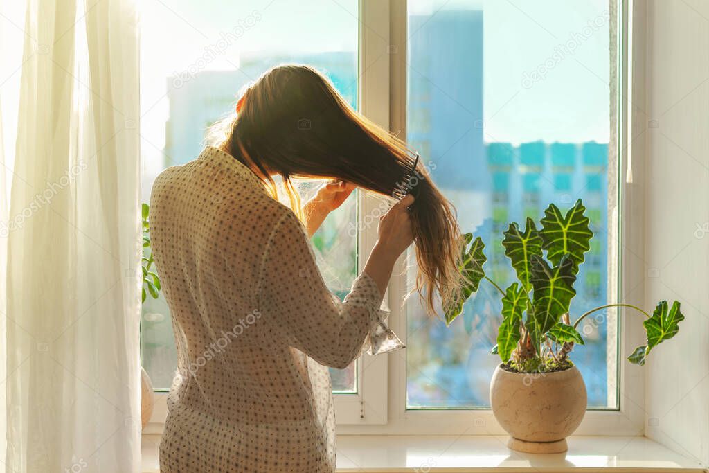 Woman in a transparent shirt brushing long brown hair in front of a window in a sunbeam. Morning beauty routine, lifestyle. Indoor plant on the windowsill. City building facade silhouette