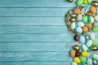 Saten kurdeleli çikolata yumurtaları. Etrafı renkli şekerlerle çevrili. Bir dal çelengi. Açık mavi ahşap zemin ve kopyalama alanı. Paskalya kutlaması için modern tebrik kartı..