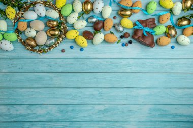 Çikolatalı tavşancık, saten kurdeleli, etrafı tatlı şekerlerle, yumurtalarla, renkli şekerlerle çevrili. Bir dal çelengi. Açık mavi ahşap zemin ve kopyalama alanı. Paskalya kutlaması için modern tebrik kartı..