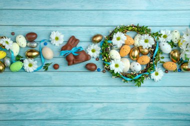 Çikolatalı tavşancık, saten kurdeleli, etrafı tatlı şekerlerle, yumurtalarla, renkli şekerlerle çevrili. Bir dal çelengi. Açık mavi ahşap zemin ve kopyalama alanı. Paskalya kutlaması için modern tebrik kartı..