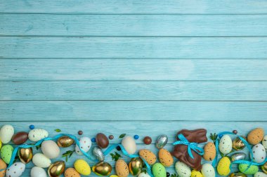 Çikolatalı tavşancık, saten kurdeleli, etrafı tatlı şekerlerle, yumurtalarla, renkli şekerlerle çevrili. Bir dal çelengi. Açık mavi ahşap zemin ve kopyalama alanı. Paskalya kutlaması için modern tebrik kartı..