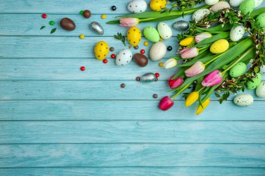 Etrafı şeker dolu buket laleler, çikolata yumurtaları, renkli şekerler. Bir dal çelengi. Açık mavi ahşap zemin ve kopyalama alanı. Paskalya kutlaması için modern tebrik kartı..