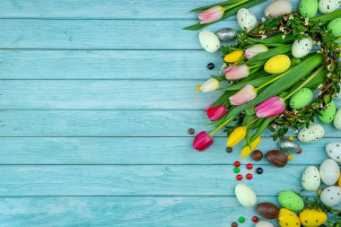 Etrafı tatlı ikramlarla, yumurta parçalarıyla, renkli şekerlerle çevrili laleler. Bir dal çelengi. Açık mavi ahşap zemin ve kopyalama alanı. Paskalya kutlaması için modern tebrik kartı..