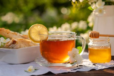 Çay ve limonlu cam bardak, ballı sürahi, hamur işleri, kruvasan, çiçek açan çalıların arasında, ahşap bir masa üzerinde bir buket yasemin dalası bulunan beyaz metal sürahi. Açık hava, piknik, brunch, gün doğumu, gün batımı. Kısa DOF ile seçili odak. Görüntüyü kapat.