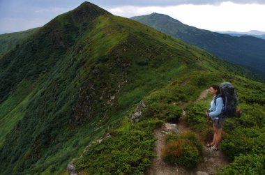 Sırt çantalı genç bir kadın bir uçurumun kenarında duruyor ve dağ manzarasına bakıyor. Turizm etkinliği.