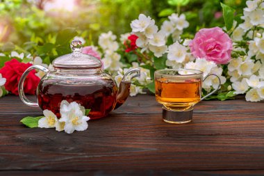 Buharda pişirilmiş bir çaydanlık ve çiçek açan gül ve yasemin çalılarının arasında zarif bir cam bardak. Açık hava, piknik, kahvaltı. Yeşil yaprakların arkaplanı bulanık.