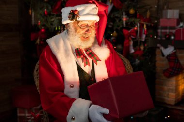 Noel Baba hediye kutusuyla şömine ve Noel ağacının yanında duruyor. Ahşap evin içindeki şenlikli iç mekan, Noel 'in neşeli ruhu. Gerçek beyaz sakallı son sınıf öğrencisi Noel Baba kostümü giyiyor..