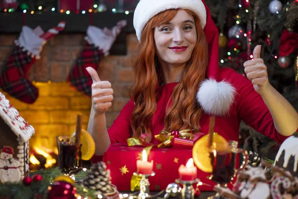 在壁炉边戴着圣诞帽的漂亮女人 在喜庆的室内挂着圣诞树 新年的气氛圣诞的精神 红头发的年轻女士为圣诞礼品盒而高兴 — 图库照片