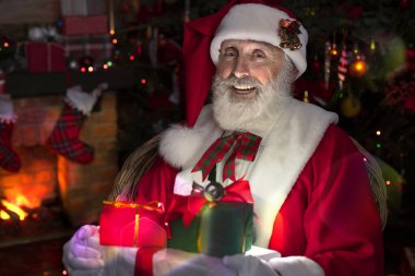 Noel Baba hediye kutusuyla şömine ve Noel ağacının yanında duruyor. Ahşap evin içindeki şenlikli iç mekan, Noel 'in neşeli ruhu. Gerçek beyaz sakallı son sınıf öğrencisi Noel Baba kostümü giyiyor..