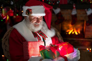 Noel Baba hediye kutusuyla şömine ve Noel ağacının yanında duruyor. Ahşap evin içindeki şenlikli iç mekan, Noel 'in neşeli ruhu. Gerçek beyaz sakallı son sınıf öğrencisi Noel Baba kostümü giyiyor..