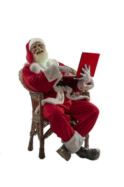 白色背景的圣诞老人被隔离了 老男演员 留着真正的白胡子 扮演圣诞老人的角色 坐在柳条长椅上 手持红色笔记本电脑 — 图库照片