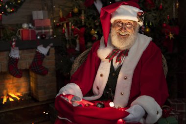 Noel Baba hediye dolu bir çantayla şömine ve Noel ağacının yanında. Ahşap evin içindeki şenlikli iç mekan, Noel 'in neşeli ruhu. Gerçek beyaz sakallı son sınıf öğrencisi Noel Baba kostümü giyiyor..